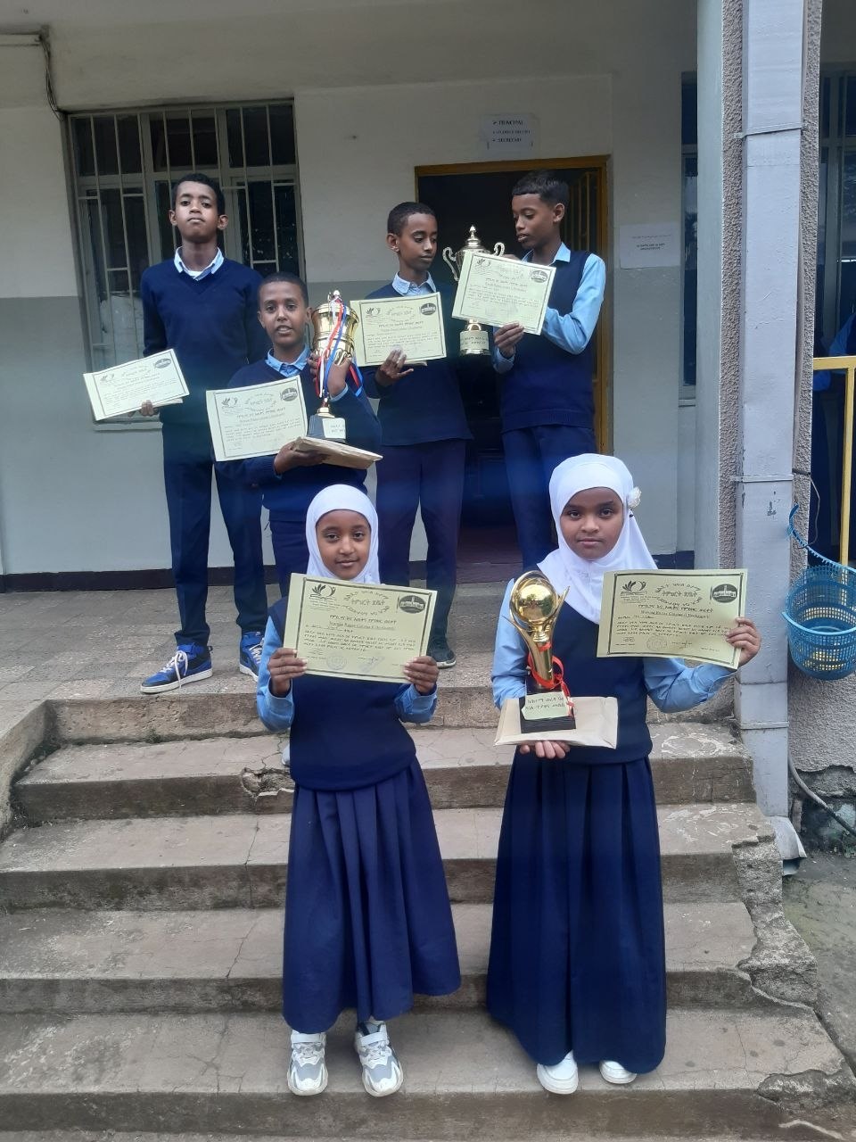 Mekanisa Abadir School students display certificates and trophies on the school steps